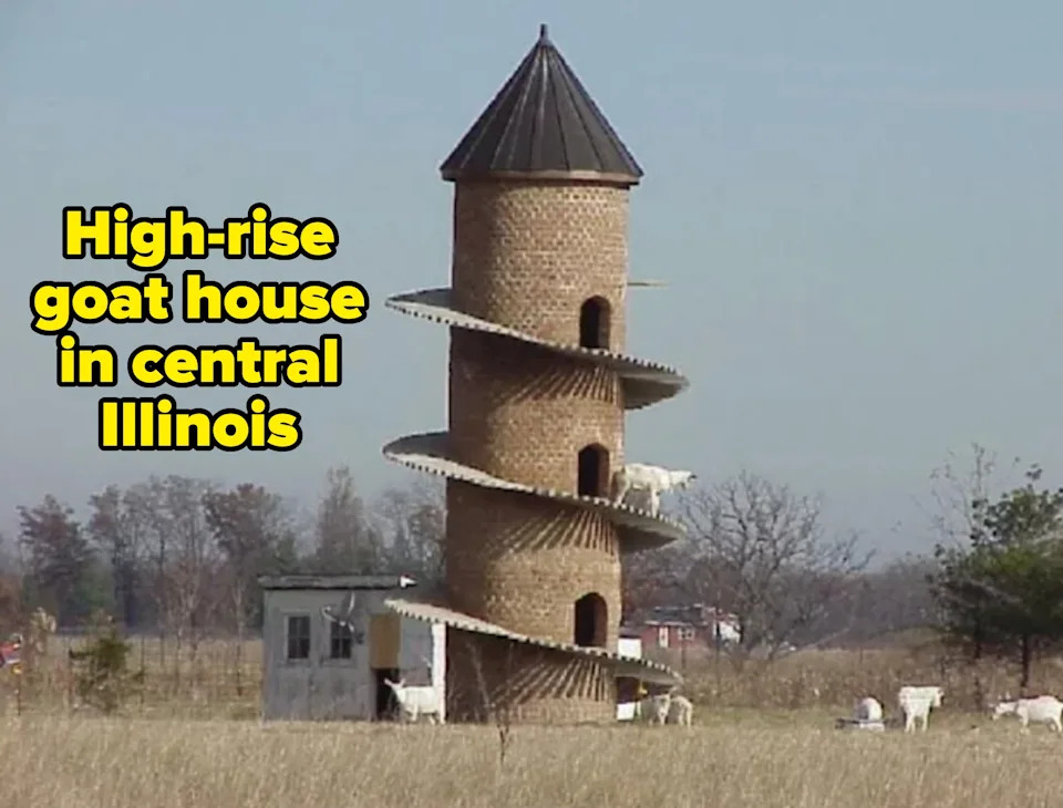 A spiral ramp winds around a tall, narrow brick tower in a field with goats walking on the ramp. A small building sits nearby