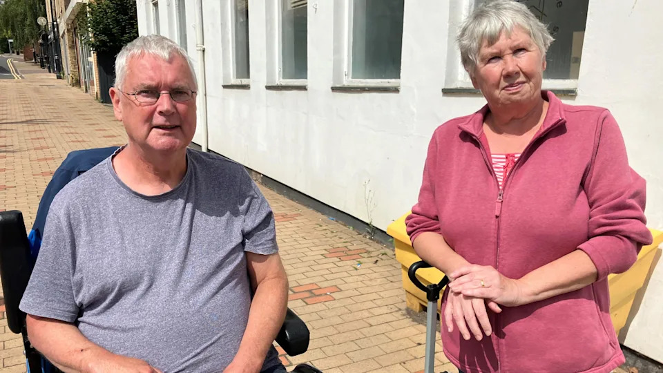A woman in a pink sweater leans on a metal pole with a handle. The man beside her, a wheelchair user, is wearing a grey t-shirt and glasses.