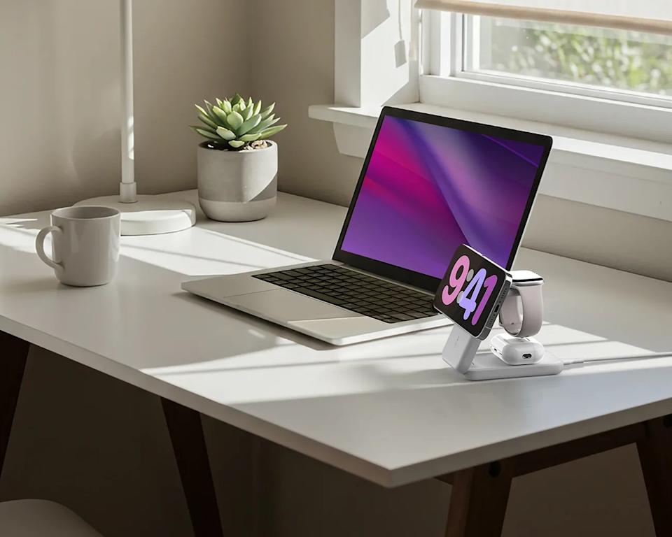 A clean white desk by a window with sunlight streaming in, featuring a silver laptop with a purple and pink screen, a white mug, a small potted succulent plant, a white desk lamp, and a white multi-device charging stand holding a smartphone, a smartwatch, and wireless earbuds.