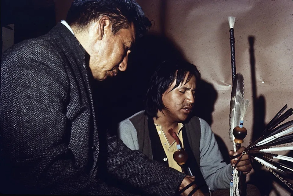 Two men, holding feathers and a small attached pot, praying with their eyes closed.