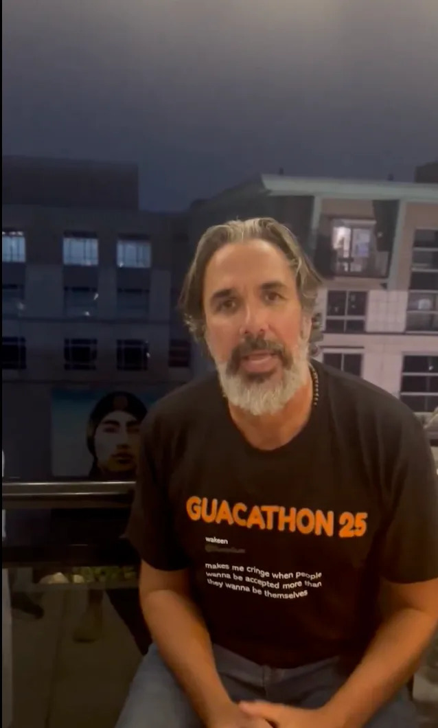 A man with long hair and a beard is sitting on a balcony wearing a "Guacathon 25" T-shirt with a quote about respecting individuality