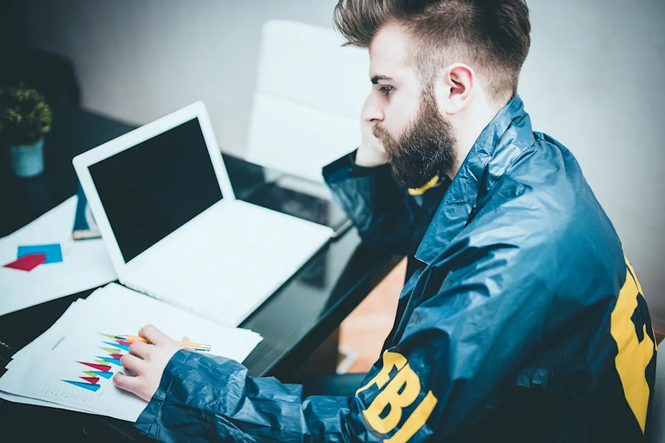 Man in FBI jacket at desk using a laptop and reviewing colorful graphs on paper, appearing focused and contemplative