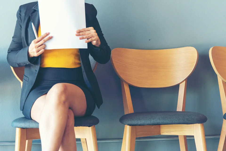 Person in a business suit sitting in a chair, holding a paper, likely at a job interview or office setting