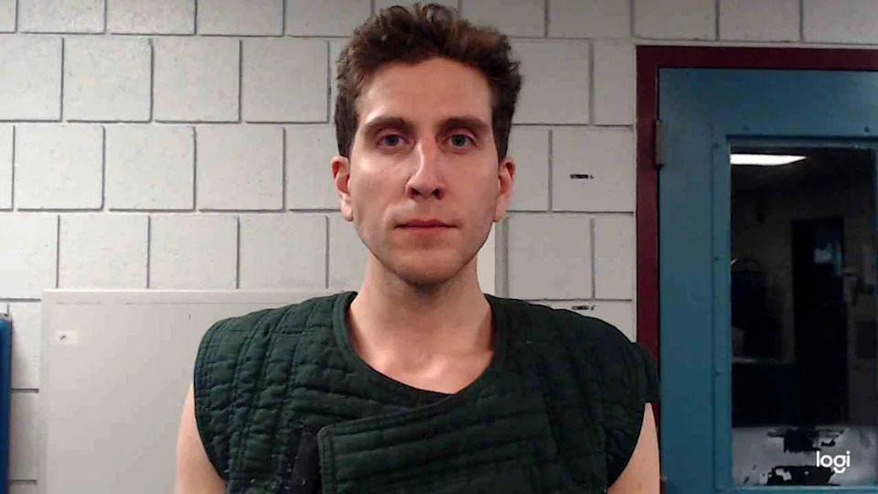 A person wearing a padded vest stands in front of a tiled wall, looking directly at the camera