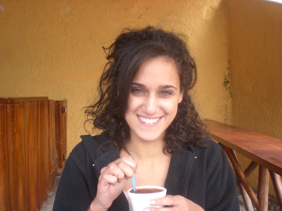 Person smiling, holding a drink with a straw in a casual setting, wearing a zipped hoodie