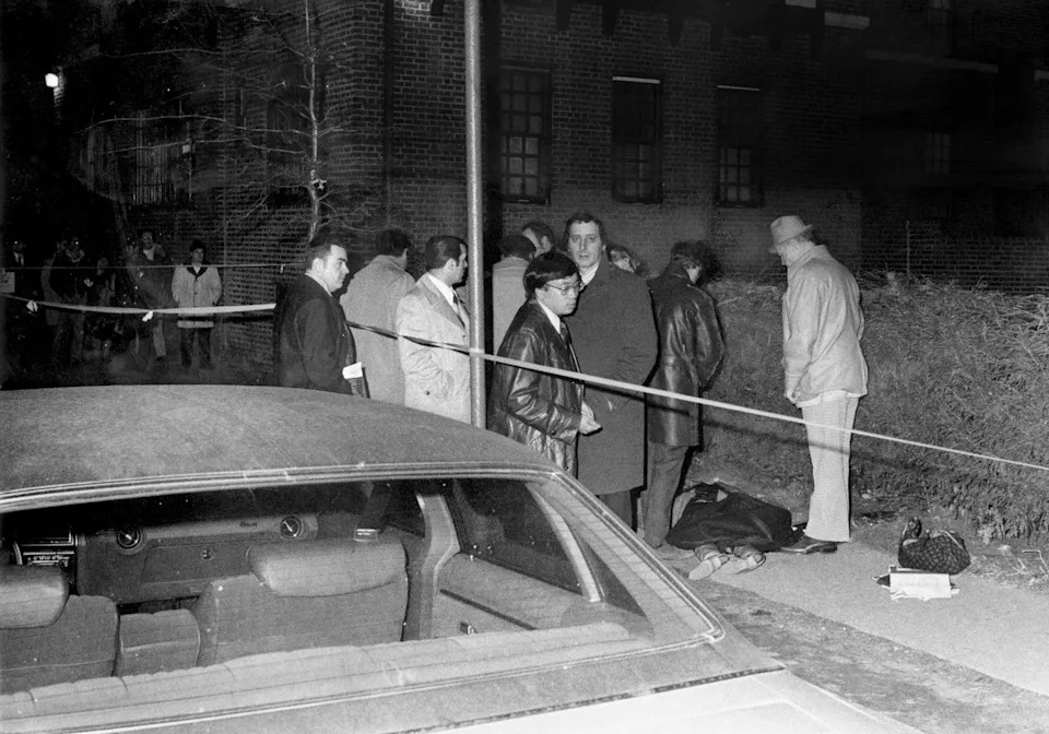a group of men stand around a sidewalk as a person lies on the ground, a car and police tape appear in the foreground