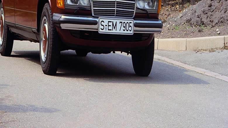 A man drives a maroon 1976-1986 Mercedes-Benz 200-280 E in front of a leafy background