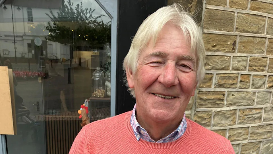A man in a salmon jumper stands in front of a window or glass door and smiles at the camera.