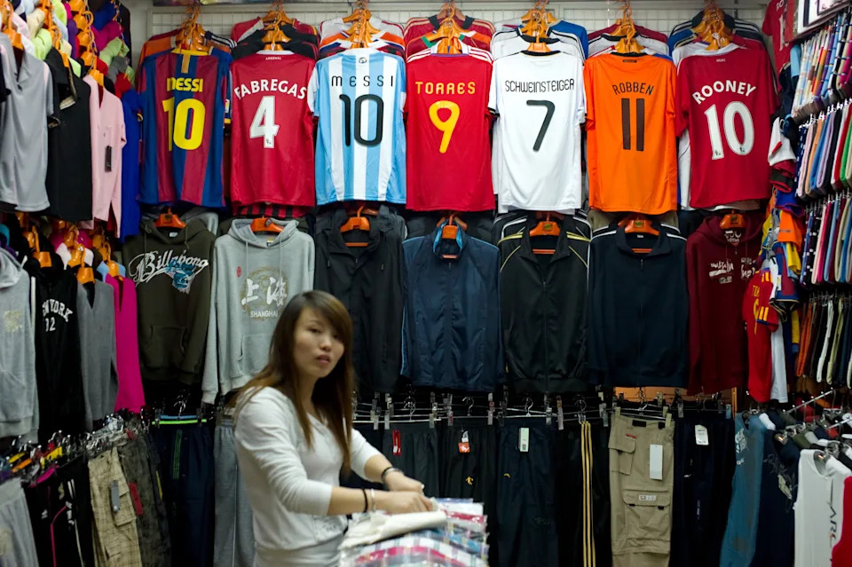Un comerciante chino coloca ropa junto a camisetas de fútbol falsas de la FIFA en un puesto de un mercado con gran cantidad de productos falsificados en Shanghái. (Foto: PHILIPPE LOPEZ/AFP vía Getty Images).