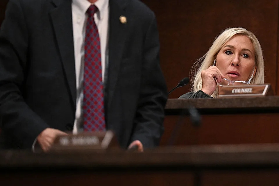 A woman is seated at a desk in a formal setting, holding glasses. A person in a suit is partially visible standing nearby