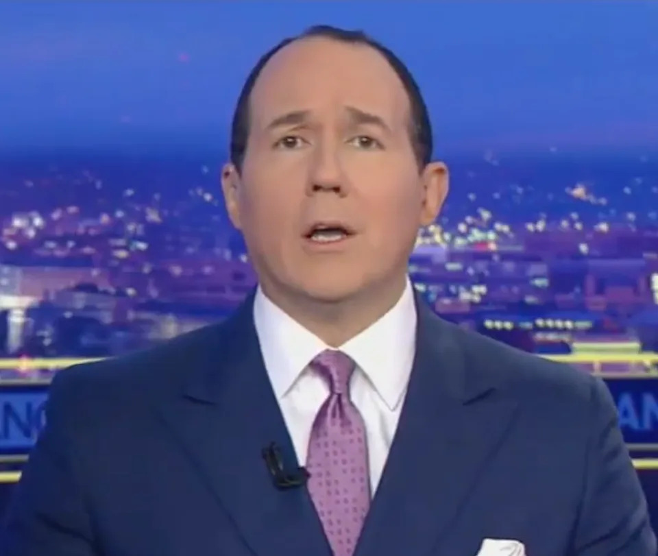 A news anchor in a suit and tie speaks during a broadcast. The cityscape is visible in the background