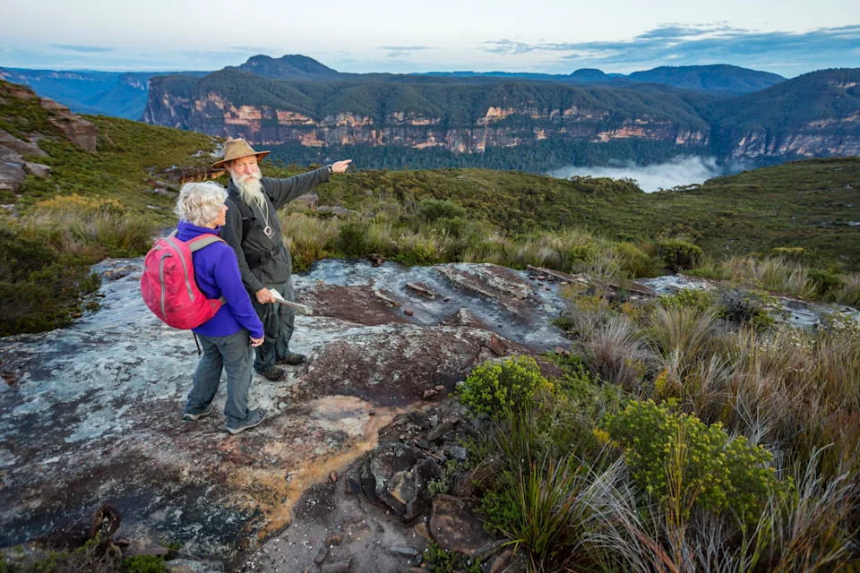 Two hikers stand on a rocky overlook, with one pointing towards distant mountains and valleys under an expansive sky