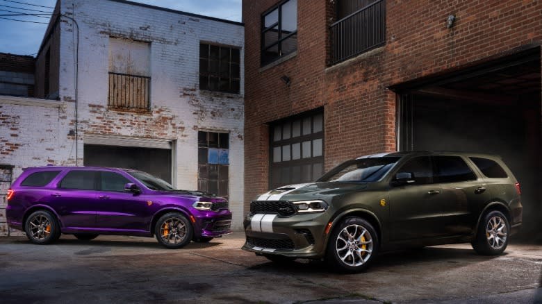 A dark green Durango Hellcat Jailbreak with silver stripes parked next to a purple Durango Hellcat Jailbreak in front of brick buildings