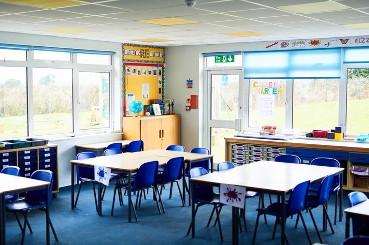 Empty classroom with blue chairs, tables, and colorful educational posters on the walls. Large windows provide natural light and view outside