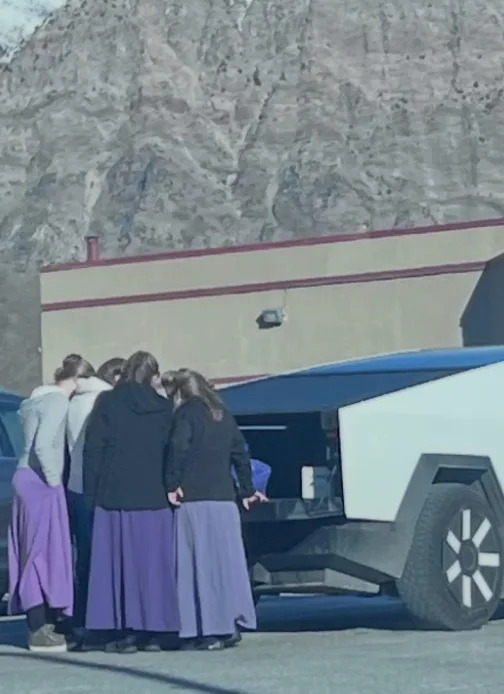 Group of people in purple skirts gather around a futuristic vehicle in a parking lot with a mountain backdrop