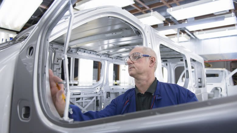 A car factory worker in safety glasses and a blue jumpsuit inspects a door hinge on a van frame