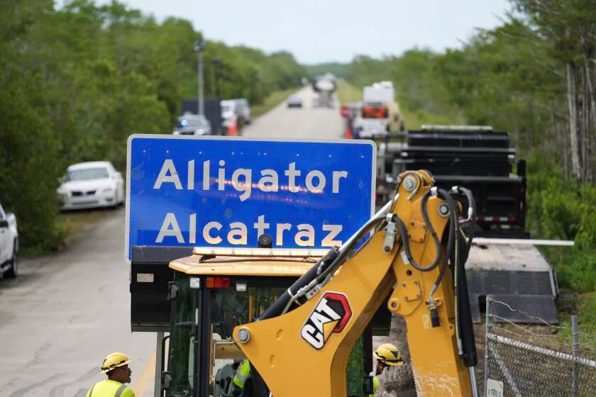 A loader holds a sign reading "Alligator Alcatraz" in its bucket as workers install it at the entrance to a new migrant detention facility at Dade-Collier Training and Transition facility, Thursday, July 3, 2025, in Ochopee, Fla. (AP Photo/Rebecca Blackwell)