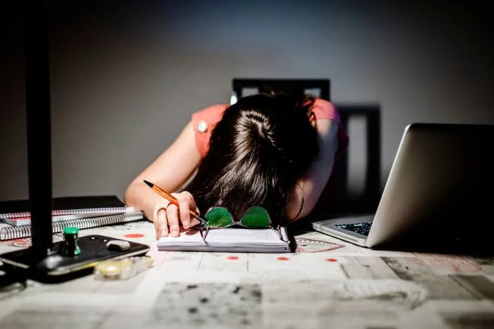 Person slumped over a table filled with papers and a laptop, head resting on folded arms, pen in hand, appearing exhausted