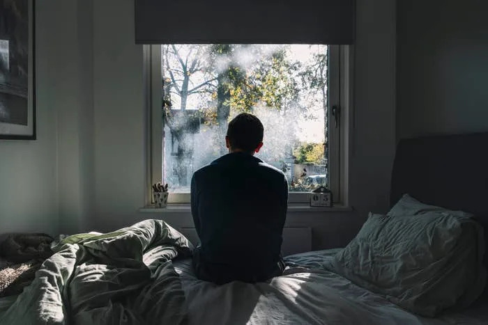 Person sits on bed facing a window, gazing outside at a sunlit view with trees. Room is softly lit, creating a reflective, tranquil atmosphere
