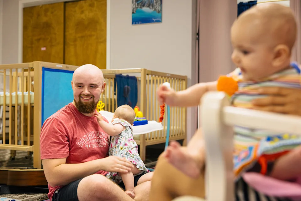 A man sits on the floor of a childcare room filled with cribs. He’s holding a baby while another sits in the foreground