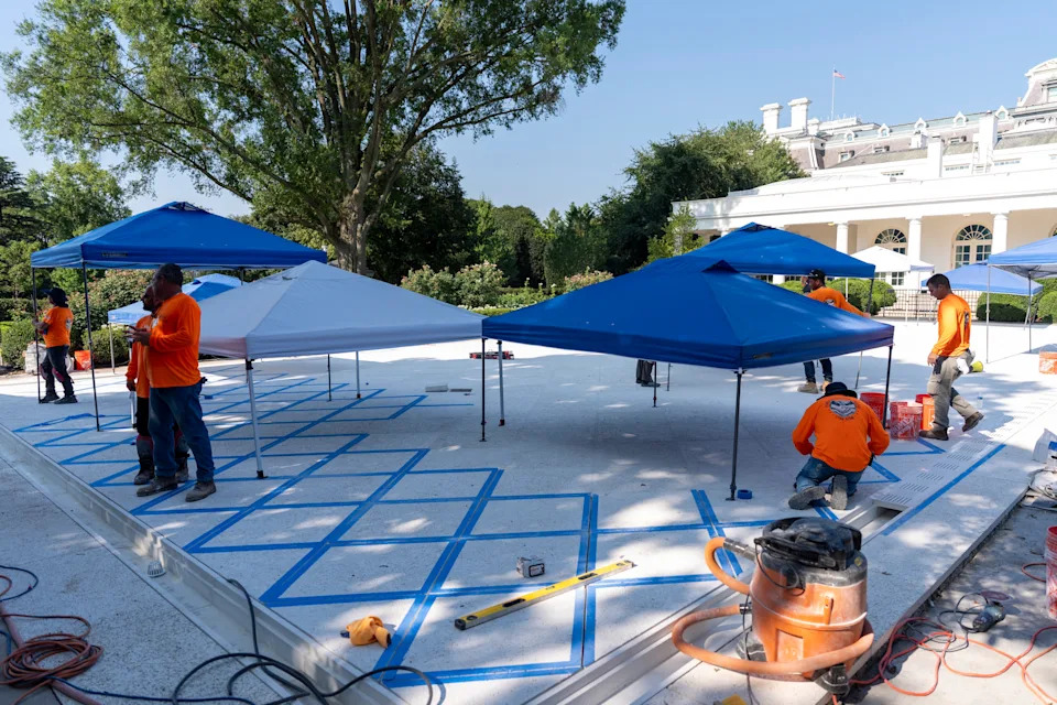 Workers setting up blue and white tents over a tiled area near a large building. Equipment and construction materials are visible