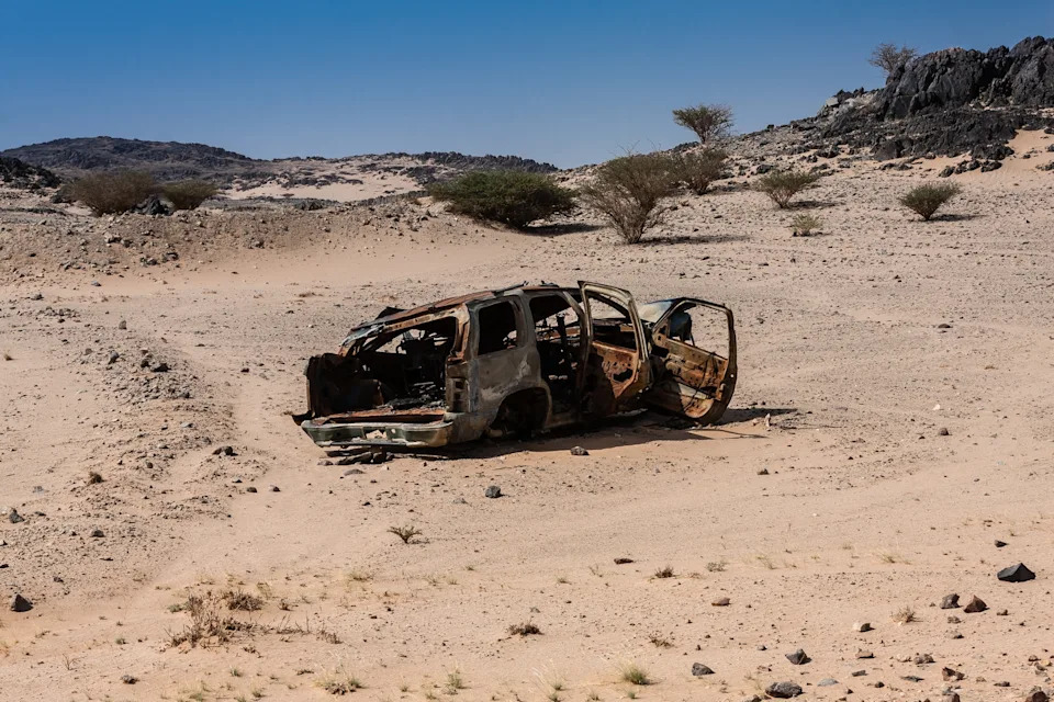 Rusted, abandoned car in a barren desert landscape with scattered shrubs and distant rocky hills