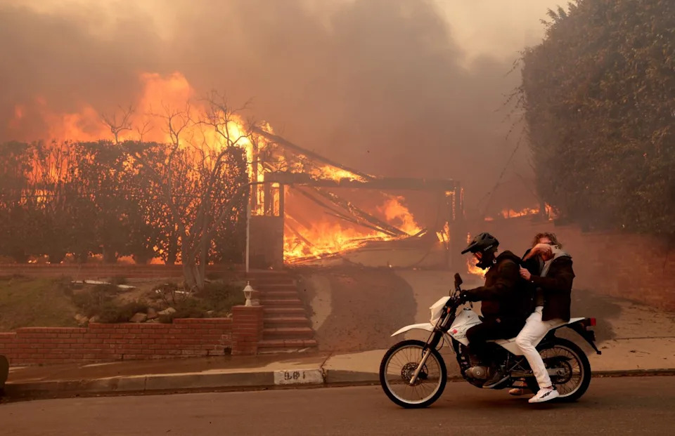 Two people ride past a burning house off Enchanted Way in the Marquez Knolls neighborhood of Pacific Palisades.