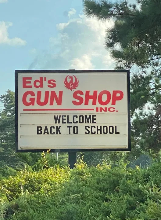 Sign reads: “Ed’s Gun Shop Inc. Welcome Back to School” against a background of trees and sky