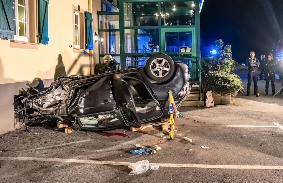 Car flipped on its side against a building at night; emergency personnel on scene