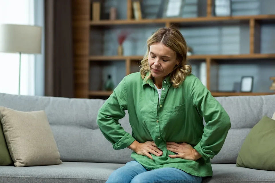 Woman leaning forward on couch with eyes closed in discomfort, hands pressing against stomach