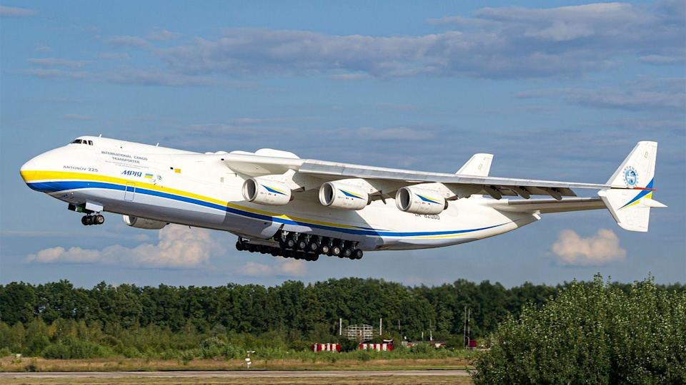 Un gran avión blanco adornado con rayas amarilla y azul a lo largo del fuselaje despega de una pista bordeada de árboles, bajo un cielo azul con unas nubes blancas. El avión tiene seis turbinas y un enorme tren de aterrizaje de 14 ruedas en el medio