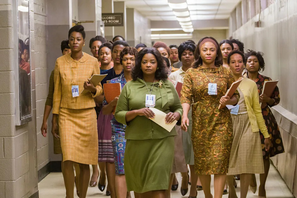 A group of women in 1960s office attire walk confidently through a hallway, holding documents