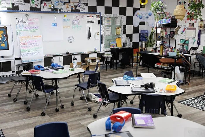 Classroom with round tables and chairs, equipped with tablets and headphones. A whiteboard displays educational materials and colorful decorations