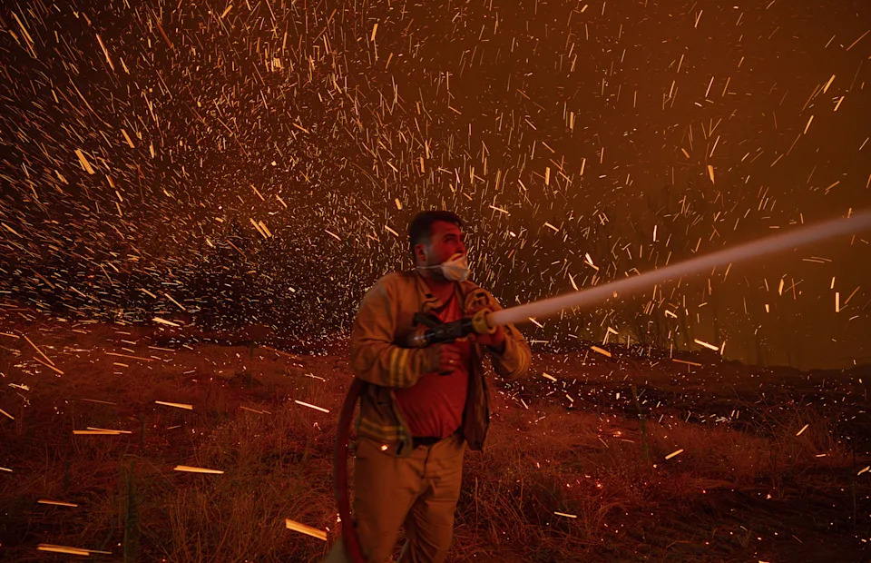 Flying embers fill the air, swirling around a firefighter spraying water from a hose.