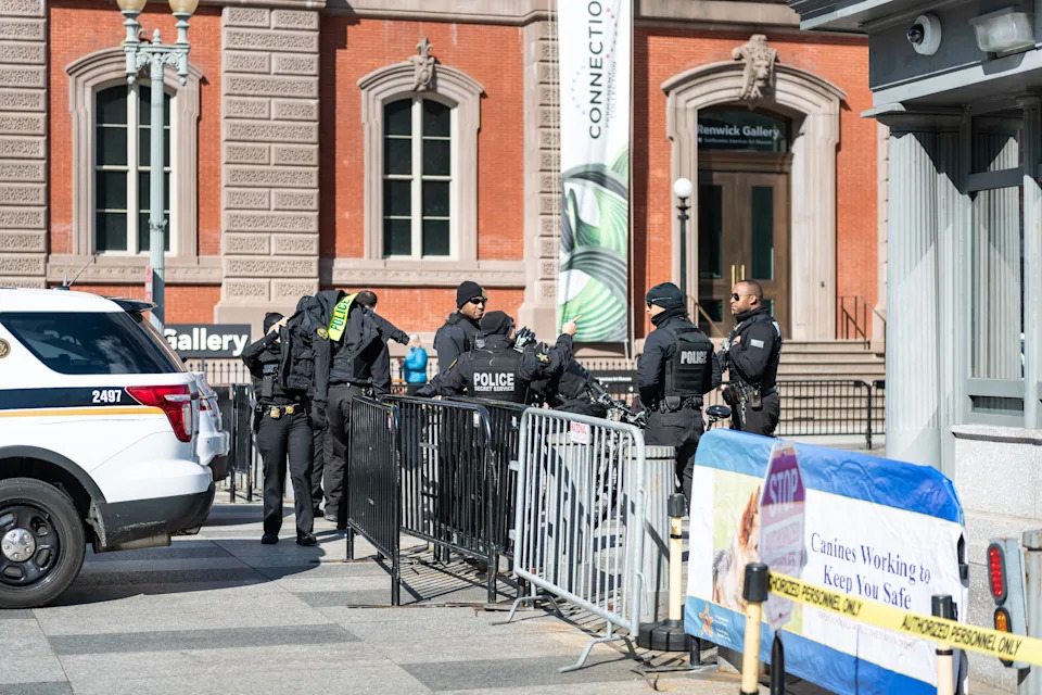 Police officers stand near barricades outside a building, engaging in serious conversation. A police vehicle is parked nearby