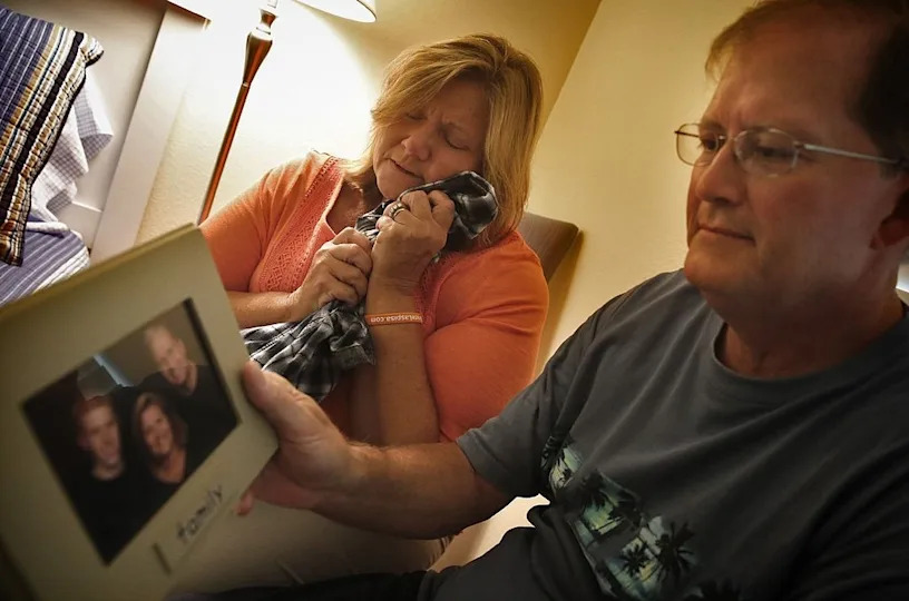 A couple looks at a family photo; the woman holds a shirt, appearing emotional, while the man observes sympathetically