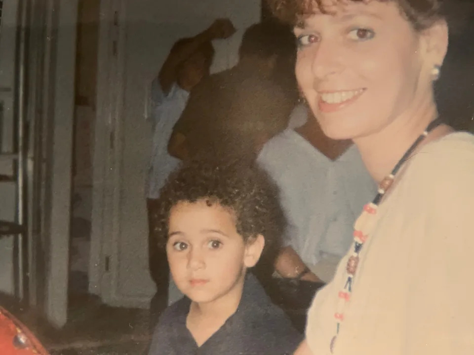A woman smiles at the camera while a young child with curly hair looks slightly to the side, seated indoors
