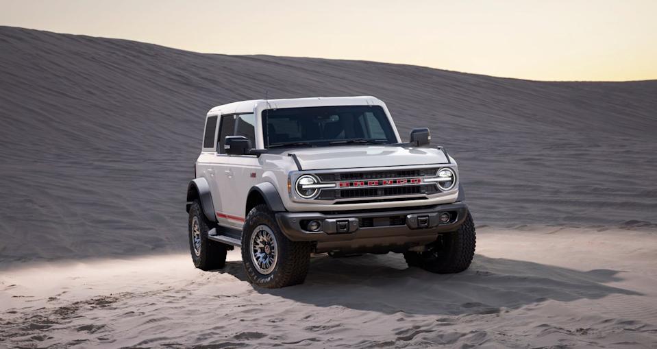 front left angle of white suv on sand with headlights on and dunes in background.