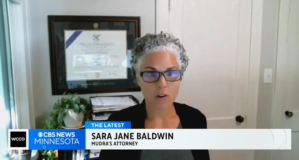 A person with short curly hair and glasses speaks in a home office with a framed certificate on the wall. TV caption reads: "Sara Jane Baldwin, Mudra's Attorney."