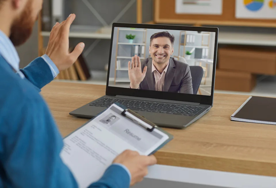 Person in a video call on a laptop with another person holding a resume. Setting suggests a virtual job interview