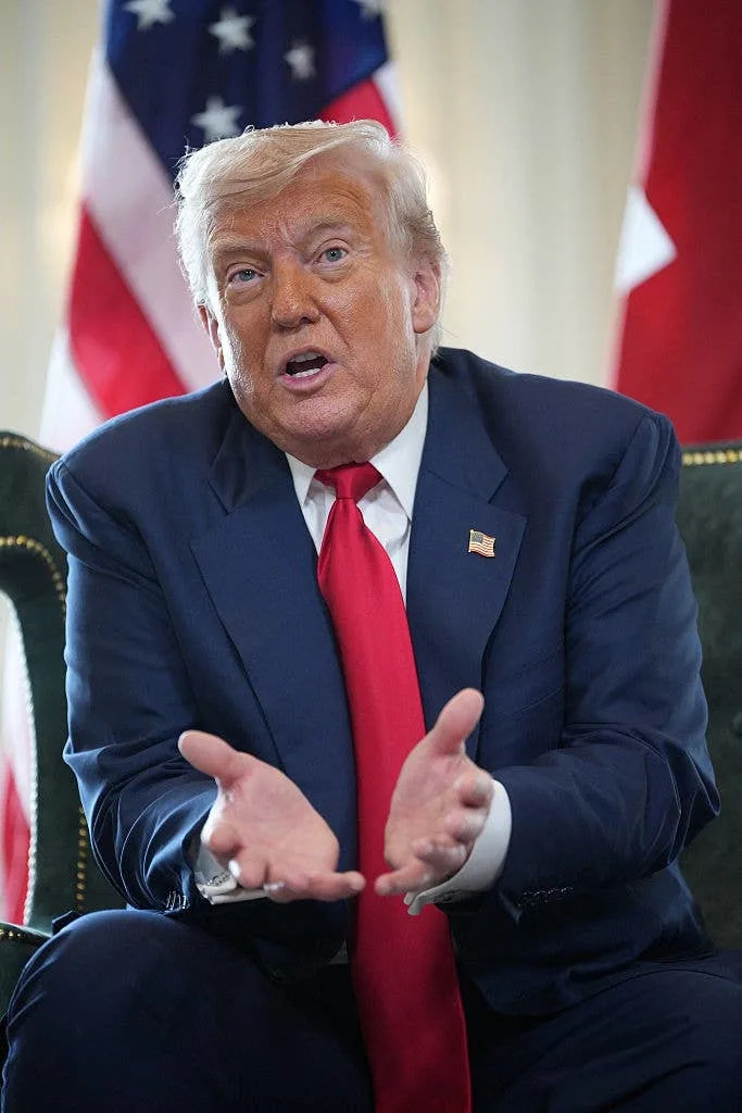 A person in a suit and red tie is gesturing with open hands during an interview or conversation, flags in the background