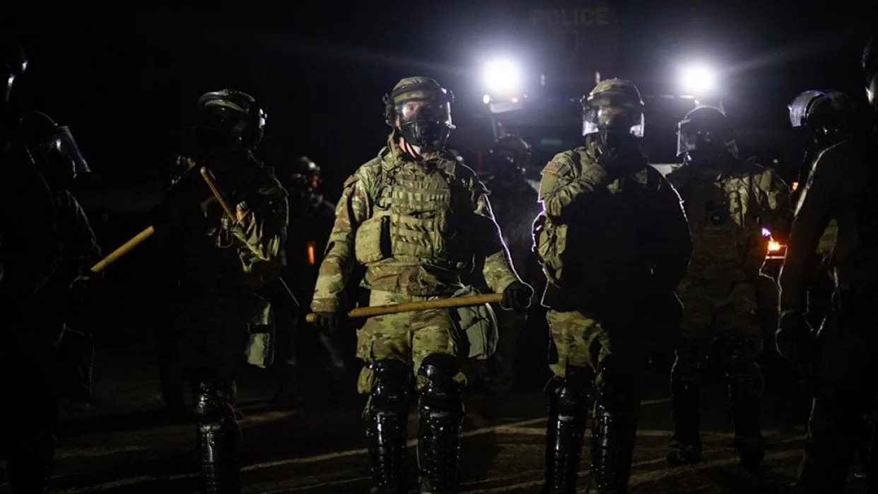 Nighttime view of a line of federal immigration agents in riot gear and face shields facing the camera near Glass House Farms facility in Camarillo, with police spotlights in background. - Larry Valenzuela // CalMatters / CatchLight Local