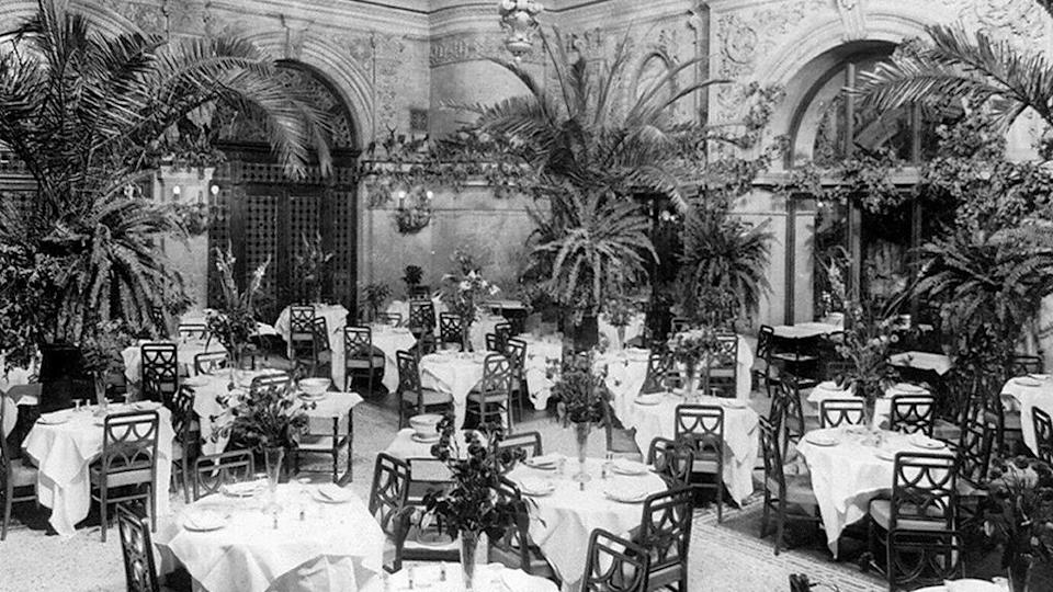 Una imagen en blanco y negro del comedor con mesas redondas vestidas con manteles blancos y un florero en el centro de cada mesa, y palmeras que decoran la sala.