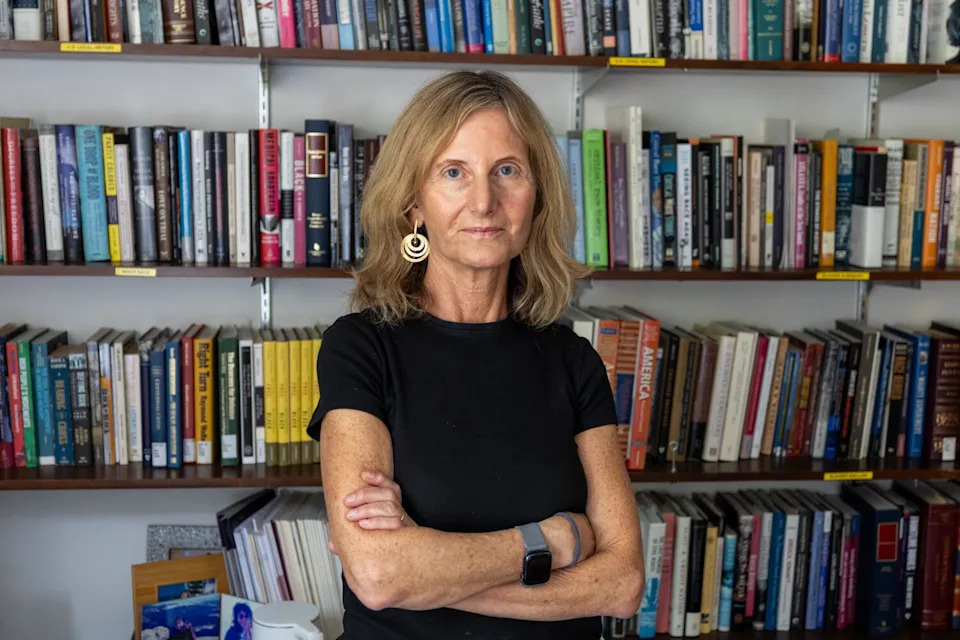 A woman stands in front of book shelves and crosses her arms.