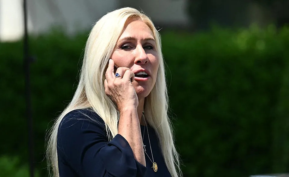 A person with long hair talks on a smartphone outdoors, wearing a dark top and a necklace