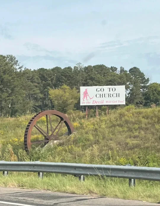 Roadside sign in a grassy area reads, "GO TO CHURCH or the Devil will get you!" with trees in the background