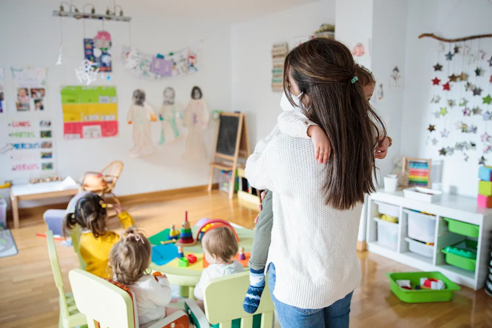 Woman holding child in a busy daycare setting with children engaged in various activities around a small table