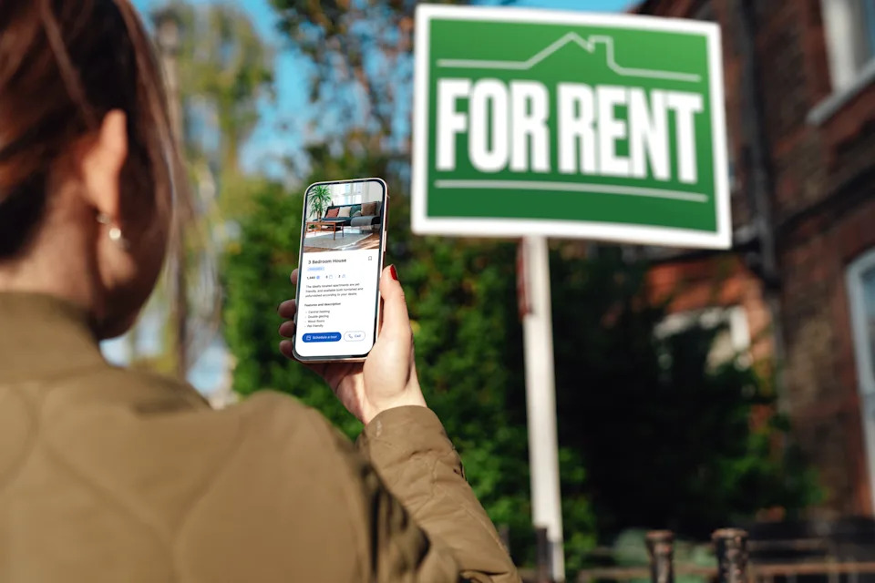 A person uses a smartphone to view a rental listing in front of a "For Rent" sign near a house