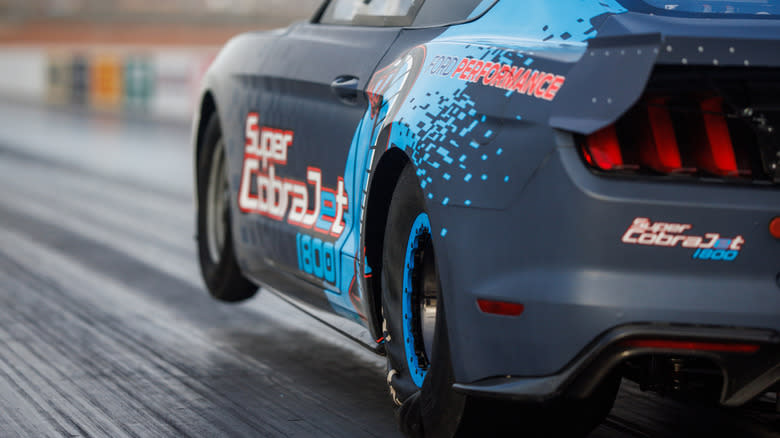 A black Ford Mustang Super Cobra Jet 1800 EV with light blue accents, at speed on a race course