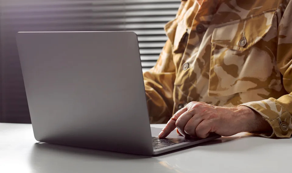 A person wearing a camouflage jacket types on a laptop at a desk, with blinds in the background